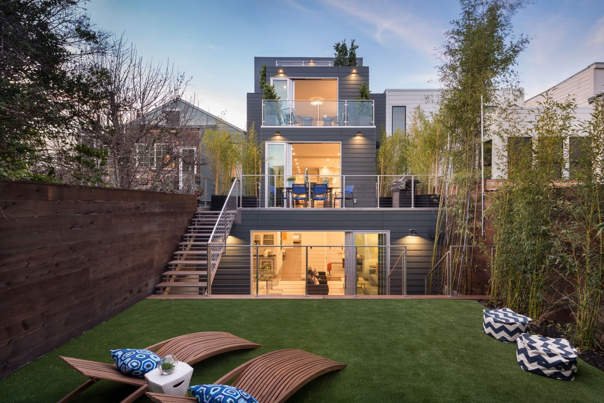 Modern three-story house with large glass windows and terraces, viewed from a backyard with wooden deck chairs and pillows on artificial grass. Tall trees and fencing surround the space.
