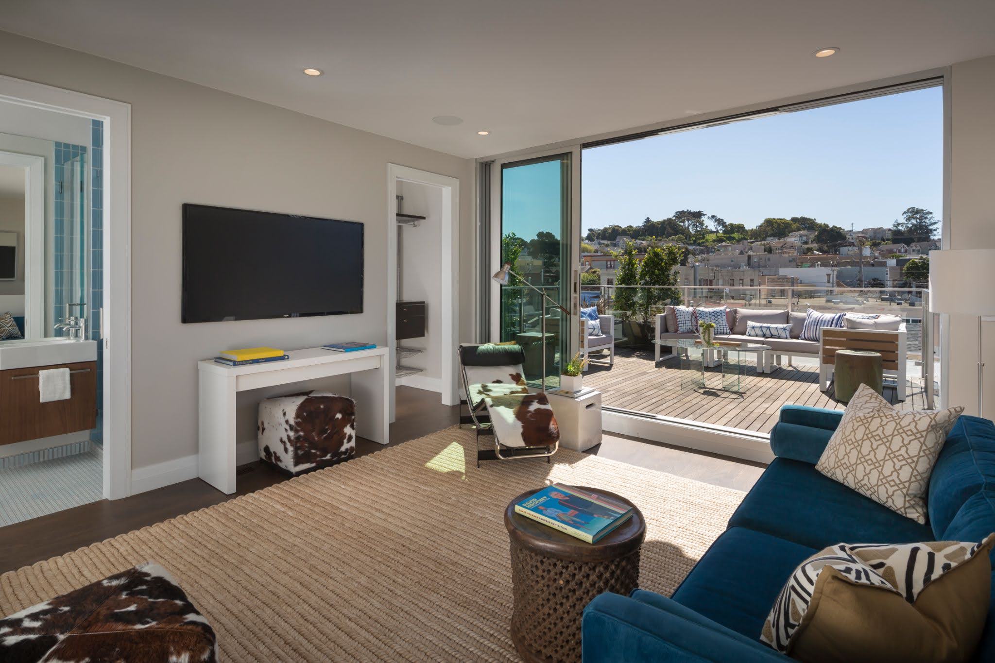 Modern living room featuring a wall-mounted TV, a blue couch, and a sliding glass door opening to a spacious balcony with outdoor seating. A bathroom is visible to the left through an open door.